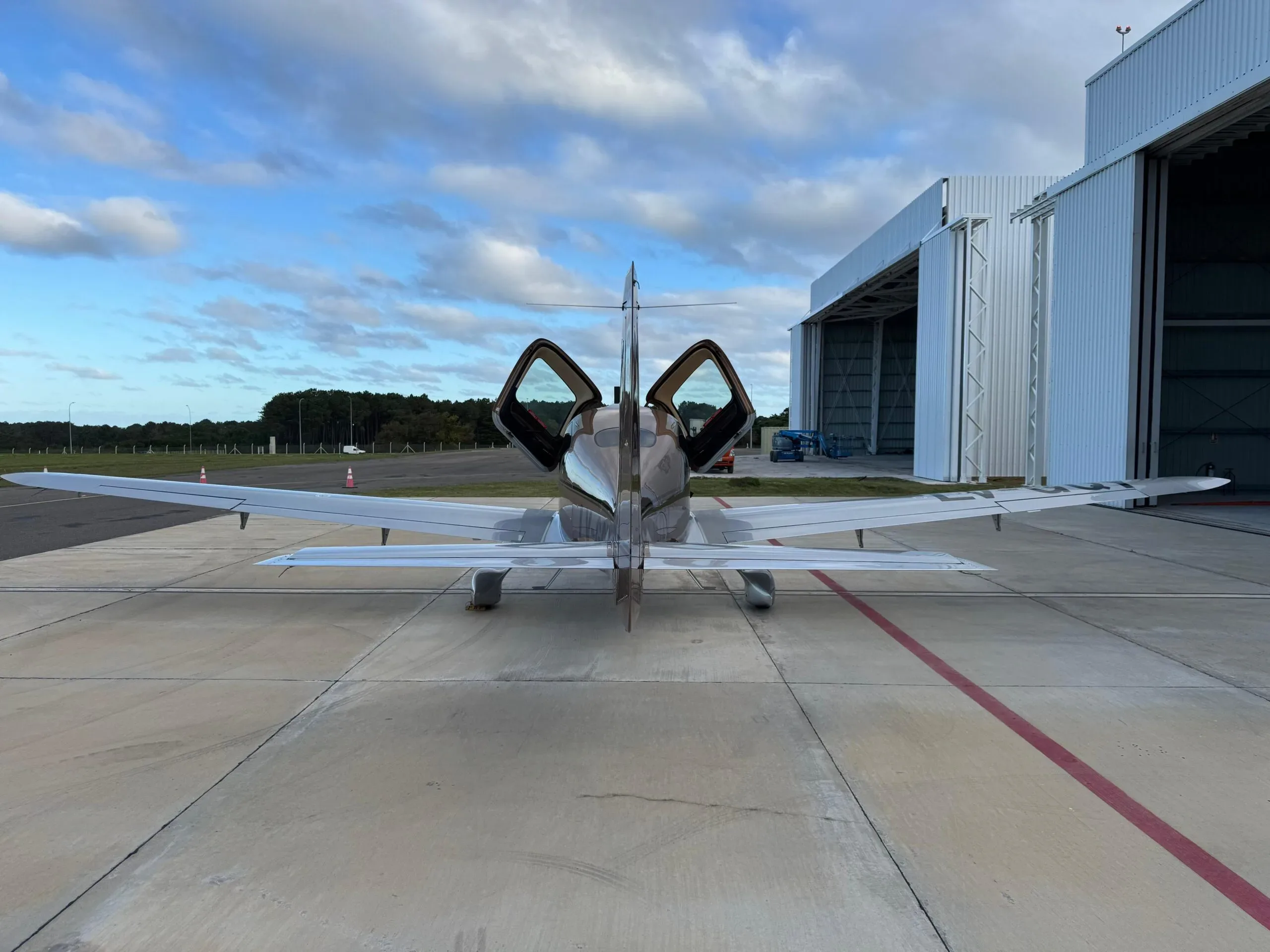A rear view of a brown and silver Cirrus SR22T aircraft with its gull-wing doors open, parked near a hangar.