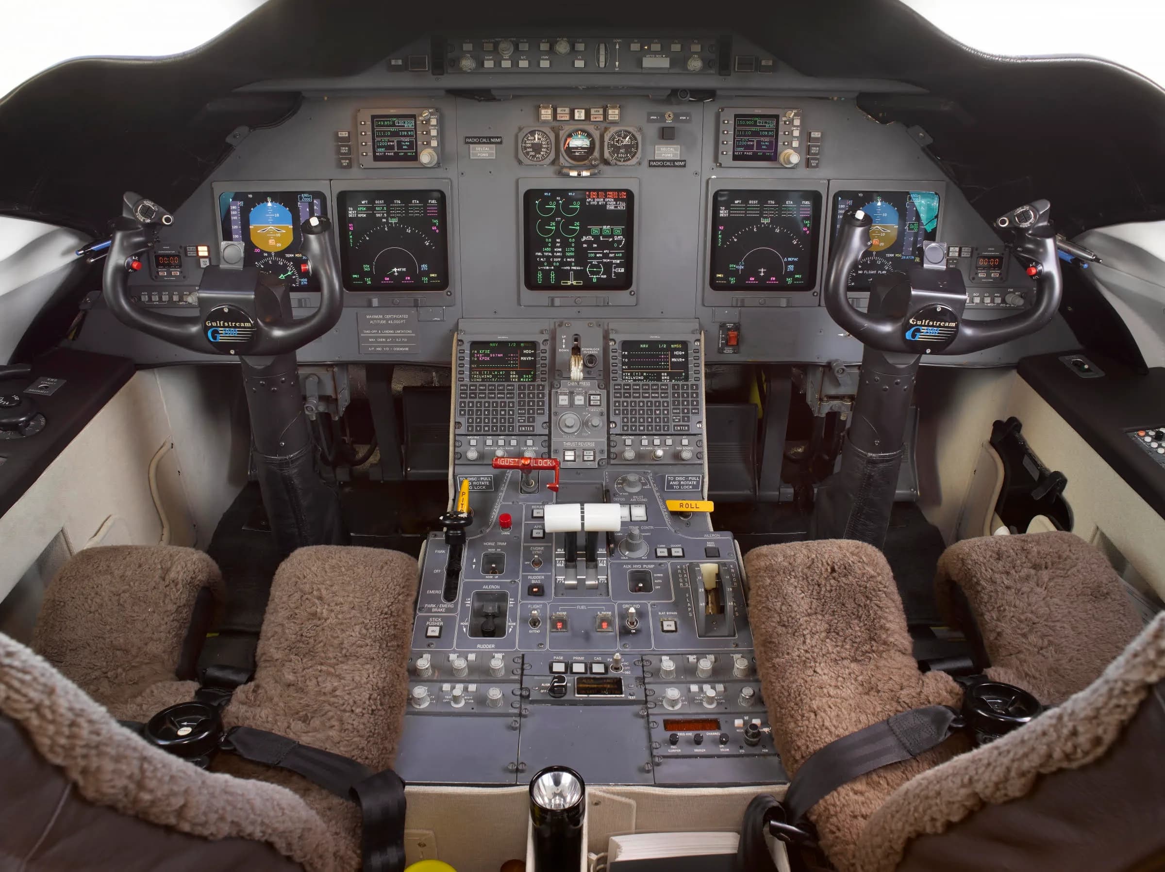 The cockpit of a Gulfstream aircraft displays multiple screens, control yokes, and a central throttle quadrant, with sheepskin seat covers.