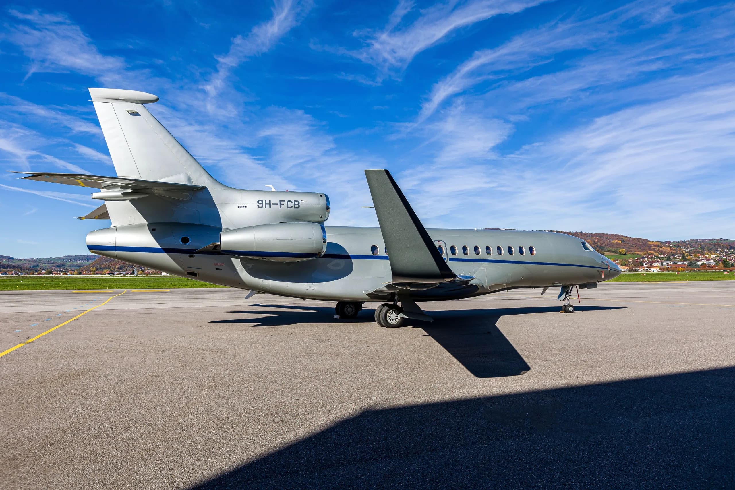 A silver Dassault Falcon 900EX business jet with blue stripes parked on an airfield, showing the rear-right side and open wing flap.