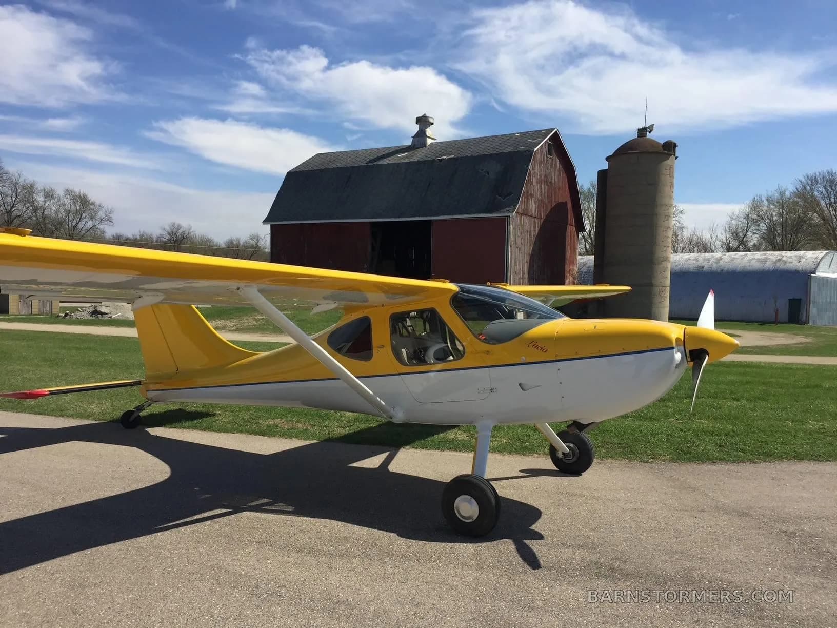 A yellow and white light sport aircraft with a blue stripe, registration N42SU, parked on a paved surface with a red barn and silos in the background.