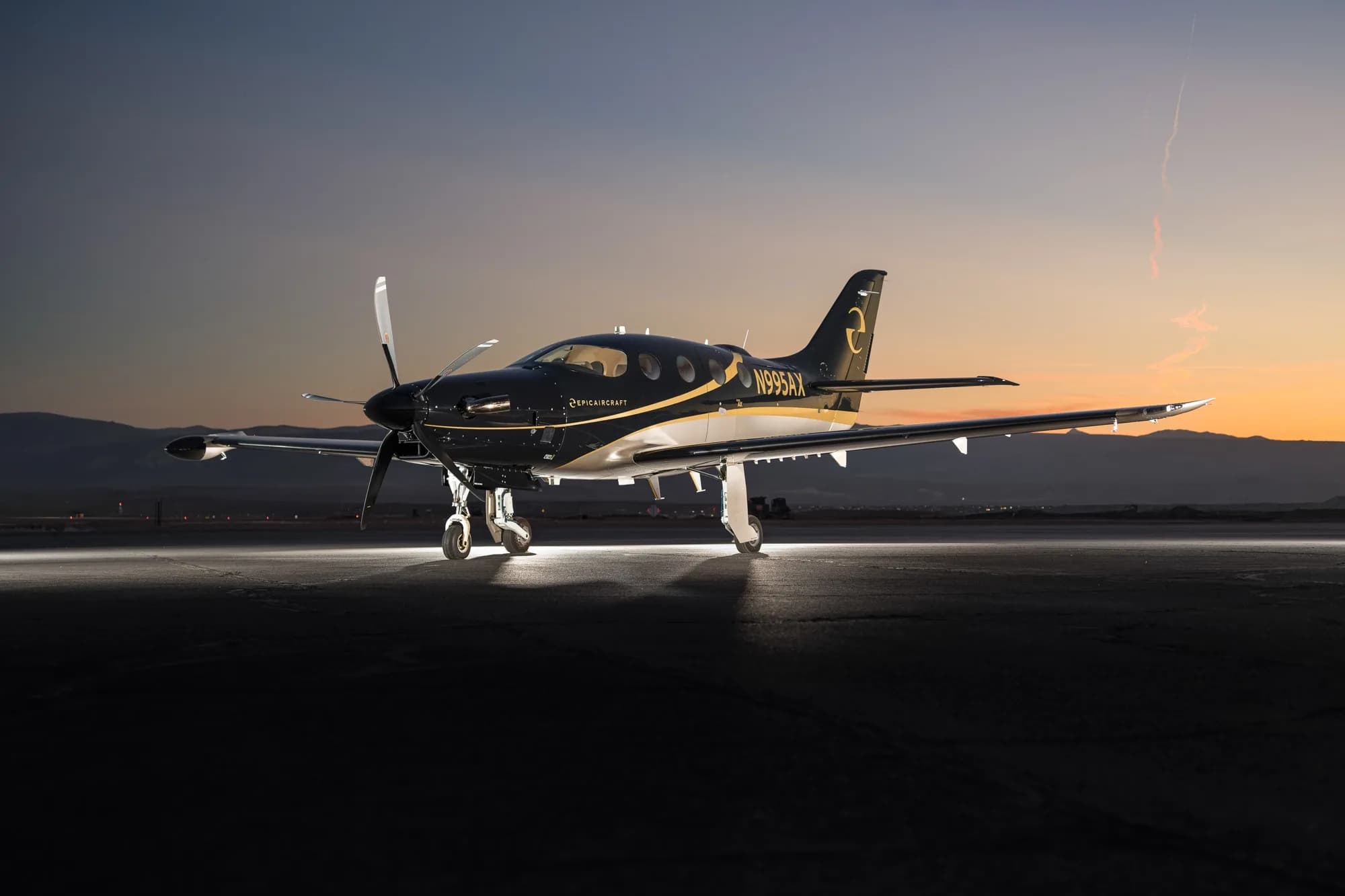 A black and gold Epic E1000 GX aircraft (N995AX) is parked on a tarmac at sunset, illuminated by ground lights.