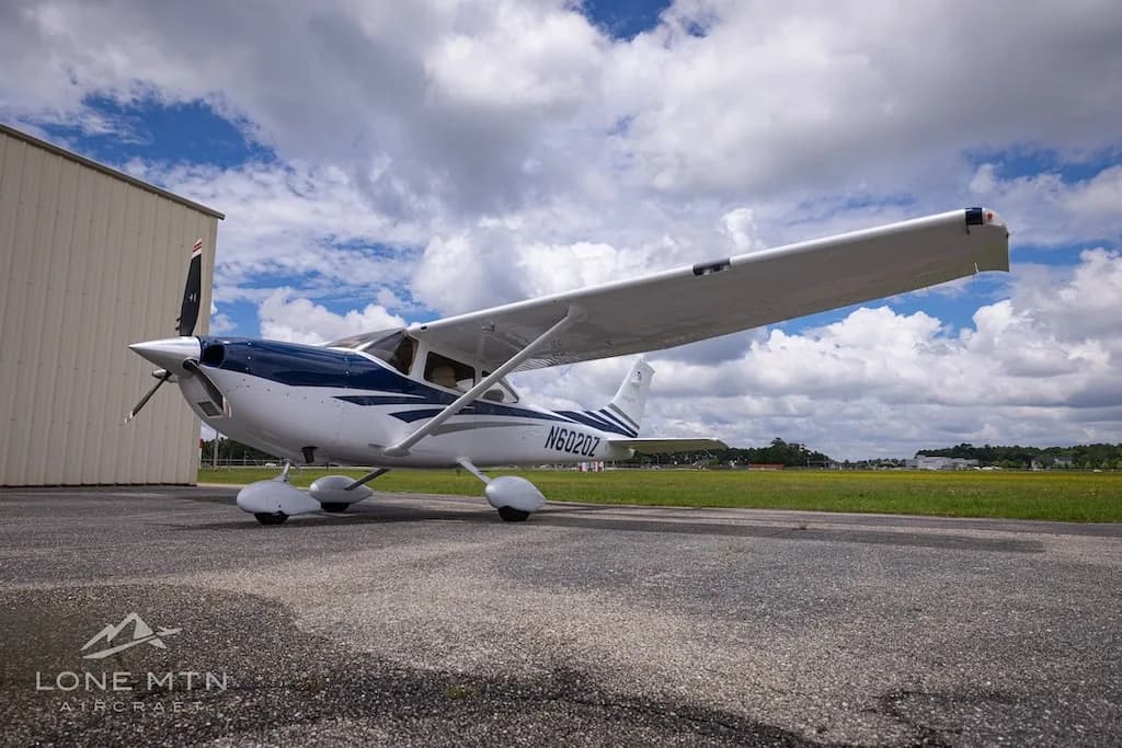 A front-left view of a white and blue Cessna Skylane TC (N6020Z) parked on an airfield next to a hangar.