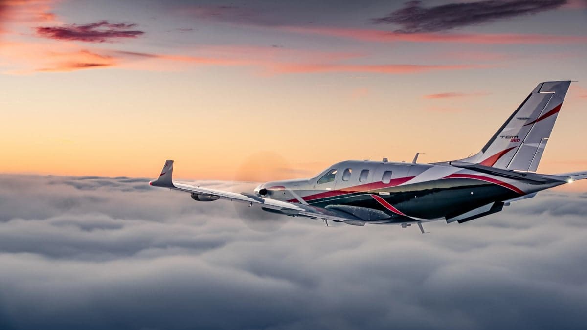 A TBM 960 aircraft with a silver, black, and red paint scheme flying above clouds at sunset.