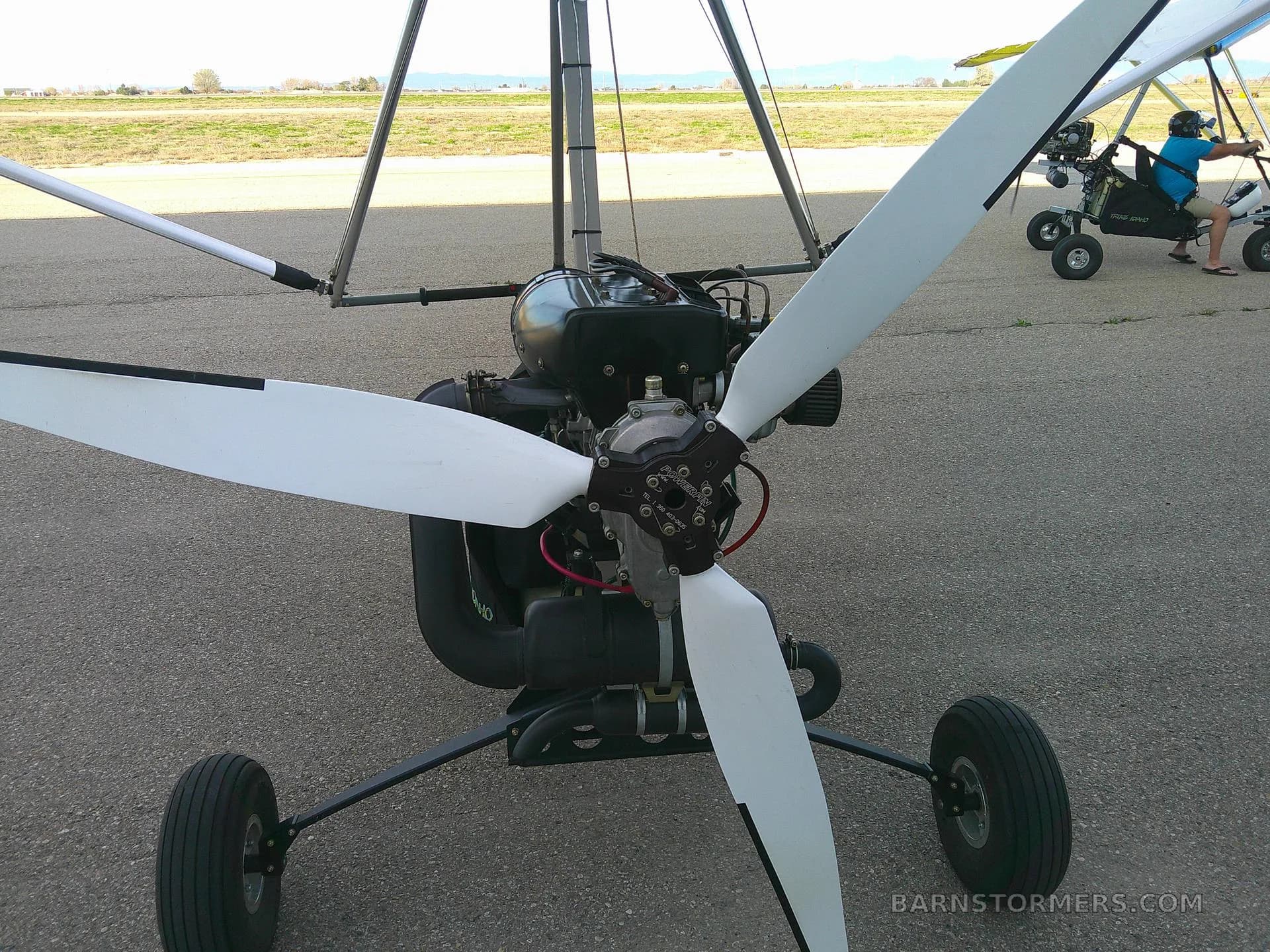 A close-up view of the black engine and white three-blade propeller of an ultralight aircraft on an asphalt surface, with another ultralight in the background.