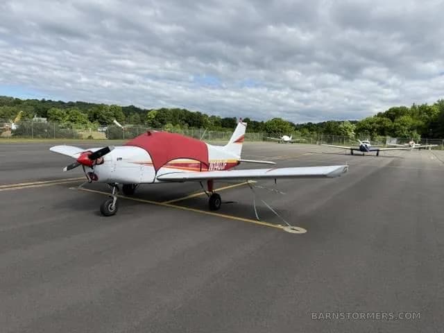 A white and red single-engine aircraft with a red engine cover parked on an asphalt apron under a cloudy sky.