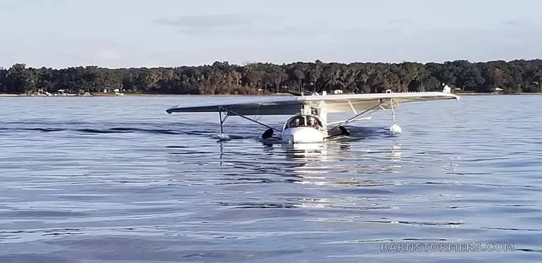 A white experimental seaplane is moving across calm water with a forested shoreline in the background.