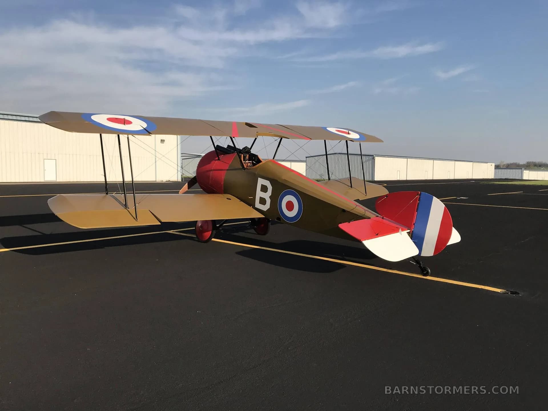 A tan and red biplane with British roundels and a French flag tail is parked on an asphalt tarmac with hangars in the distance.