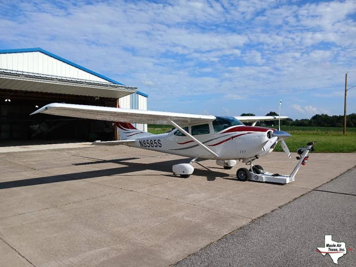 A white single-engine high-wing aircraft with red and grey stripes is parked on a concrete ramp next to an open hangar.