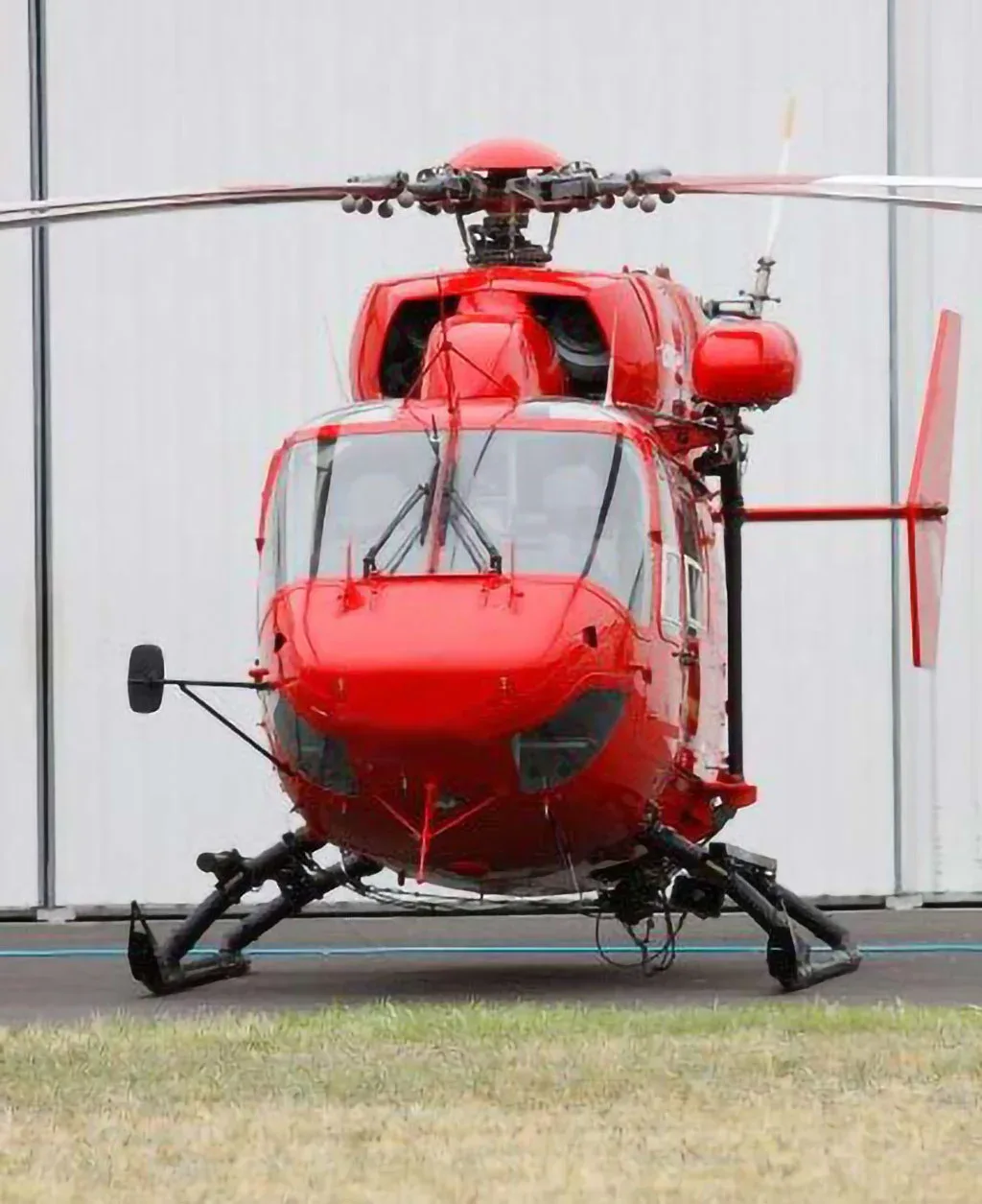 A red BK117 helicopter seen from the front, parked on a tarmac in front of a white hangar.