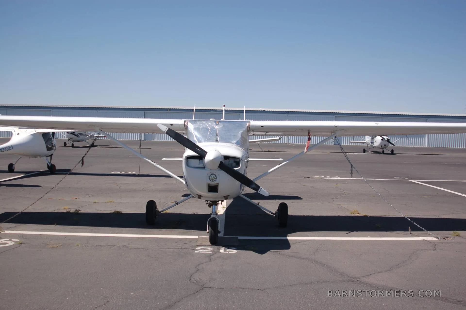 A white Cessna aircraft with a propeller is parked head-on on an airfield with hangars in the background.