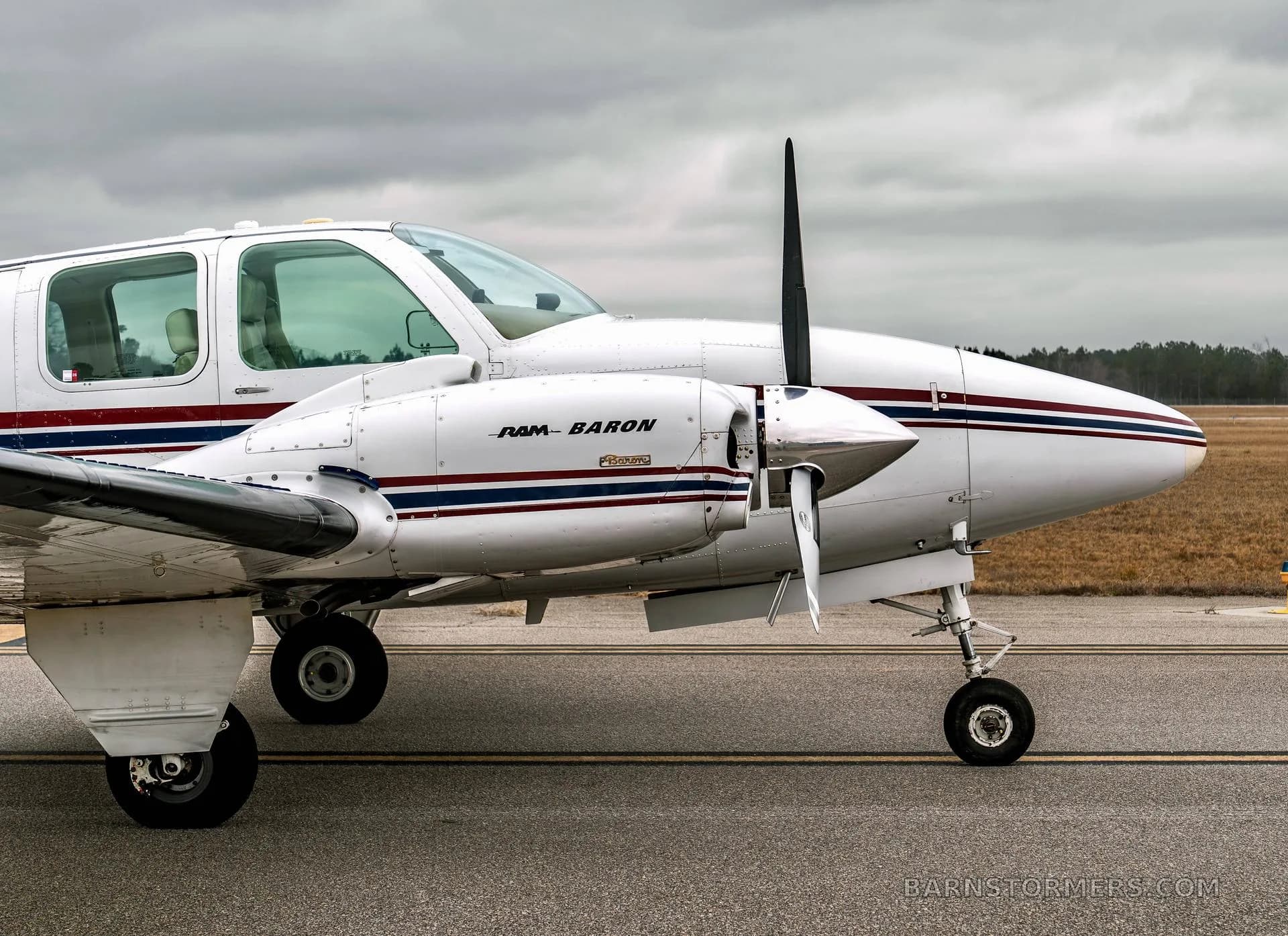 A close-up side view of the front of a white twin-engine Beechcraft Baron N158TB, showing the propeller and 'RAM BARON' text on the engine cowling.