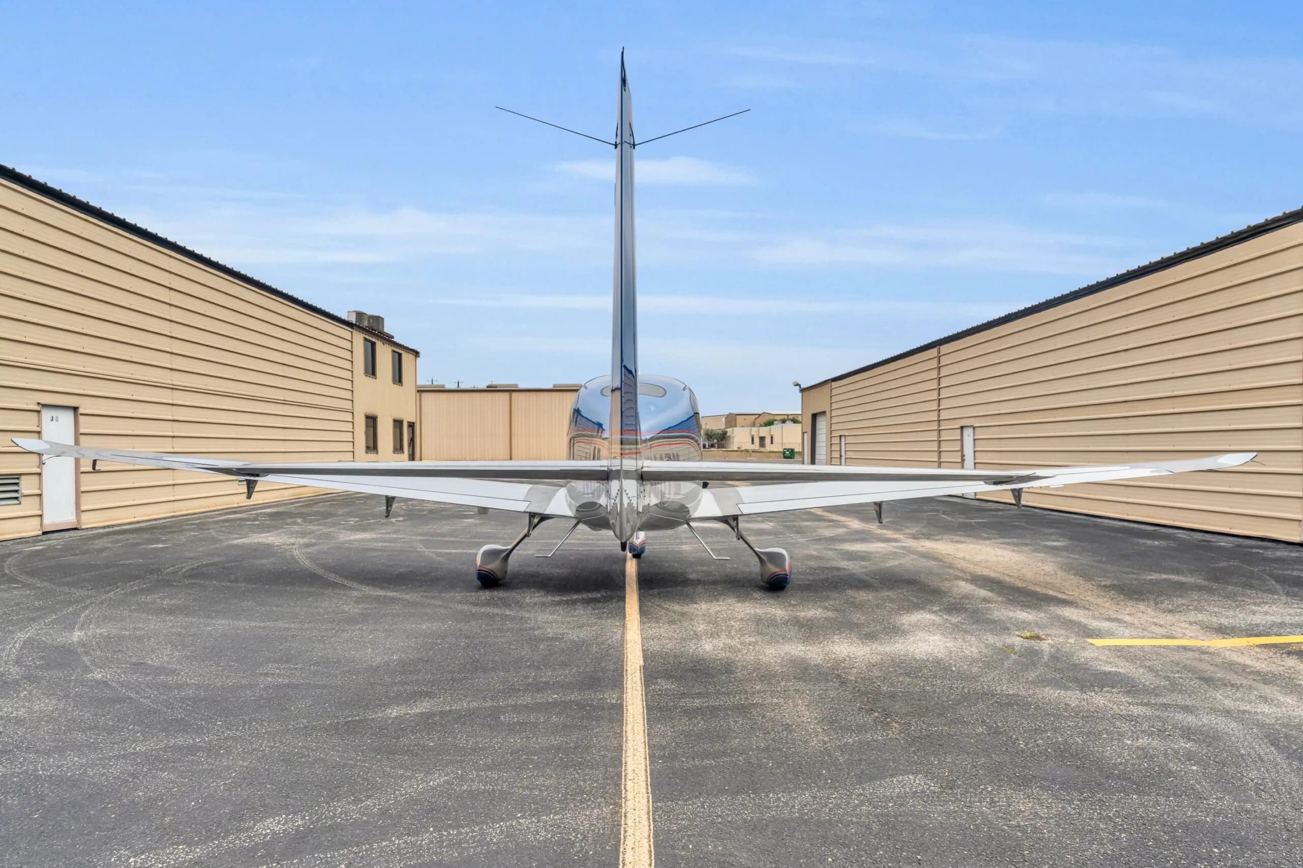 A rear view of the blue and silver Cirrus SR22T GTS aircraft N66TM, centered on an asphalt taxiway between hangars.
