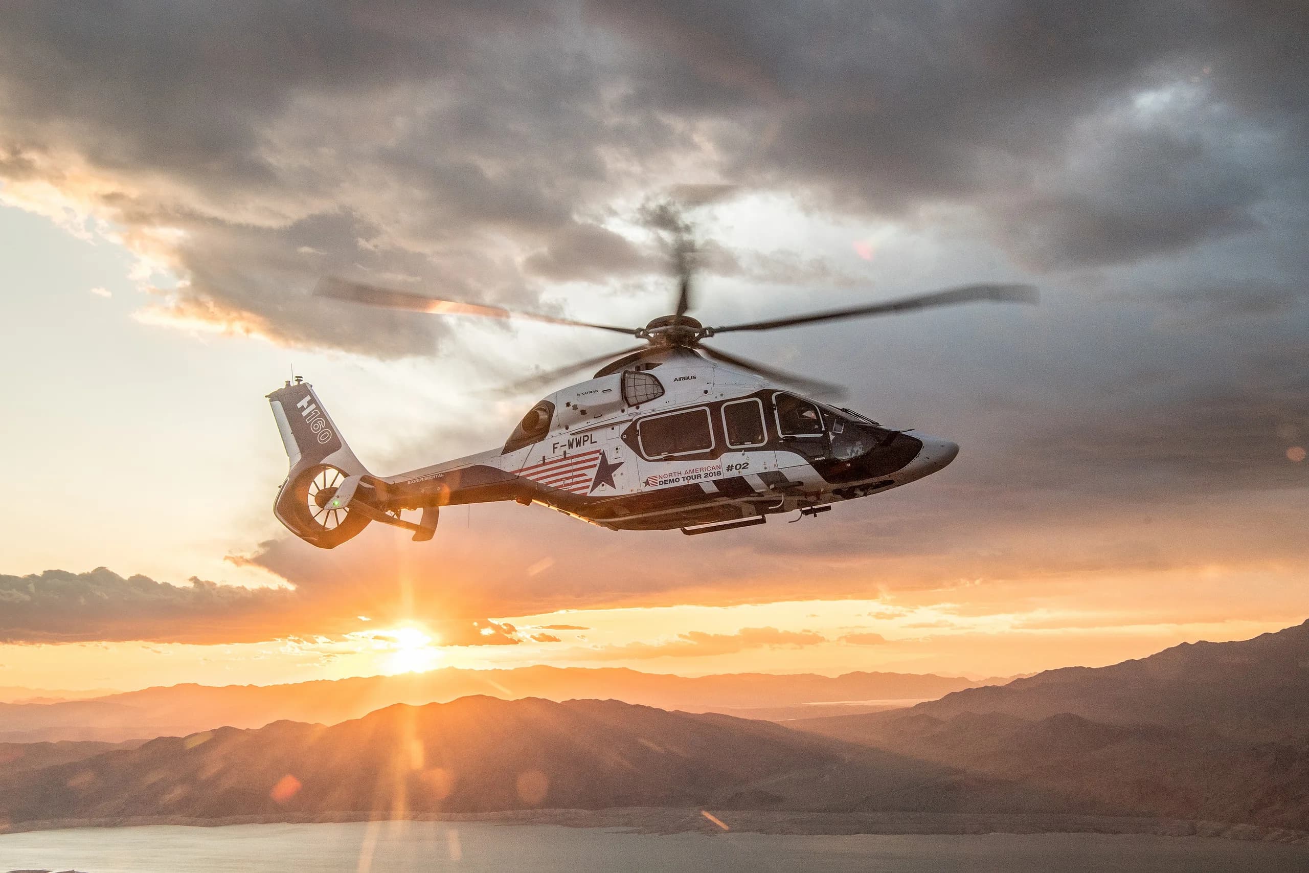 An Airbus H160 helicopter flying over a lake at sunset.