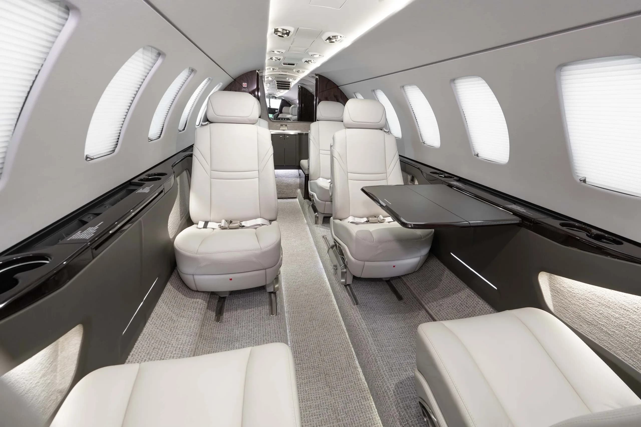 The interior cabin of a business jet with light grey leather seats and a fold-out table, looking towards the front.
