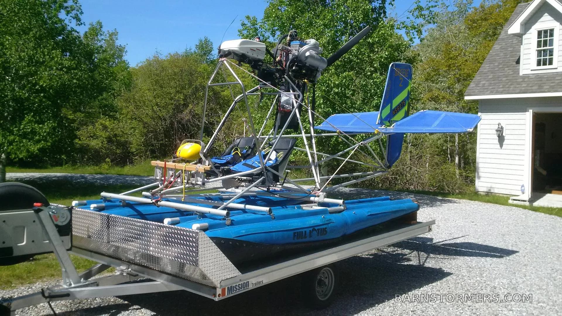 A blue ultralight seaplane with a yellow tail and exposed frame, on a trailer in a gravel driveway.