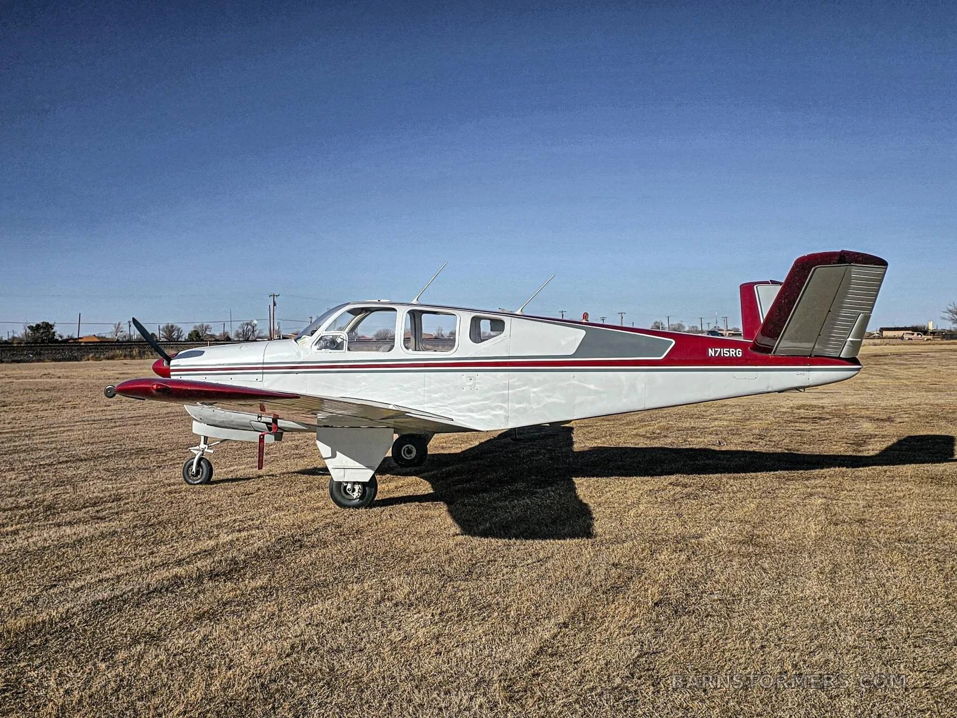 A white and red Mooney M20 series aircraft (N7158G) viewed from the side, parked on a grassy field under a clear blue sky.