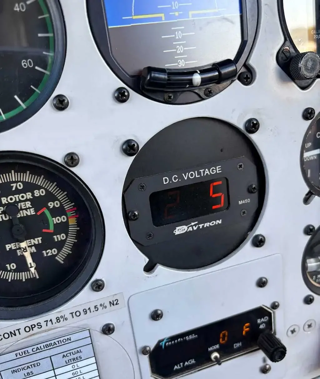 A close-up of a helicopter's instrument panel, showing a DC voltage display, rotor RPM gauge, and fuel calibration chart.