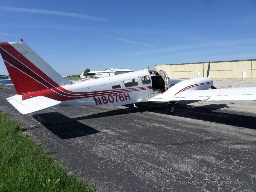 A white and red single-engine propeller aircraft N8076H with its door open, parked on an asphalt ramp next to hangars.