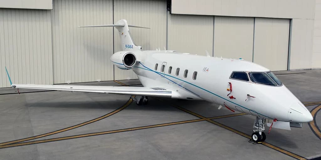 A white business jet with blue and teal stripes, registration "HK-5U5A" and "IEA", is parked on a tarmac in front of a hangar.