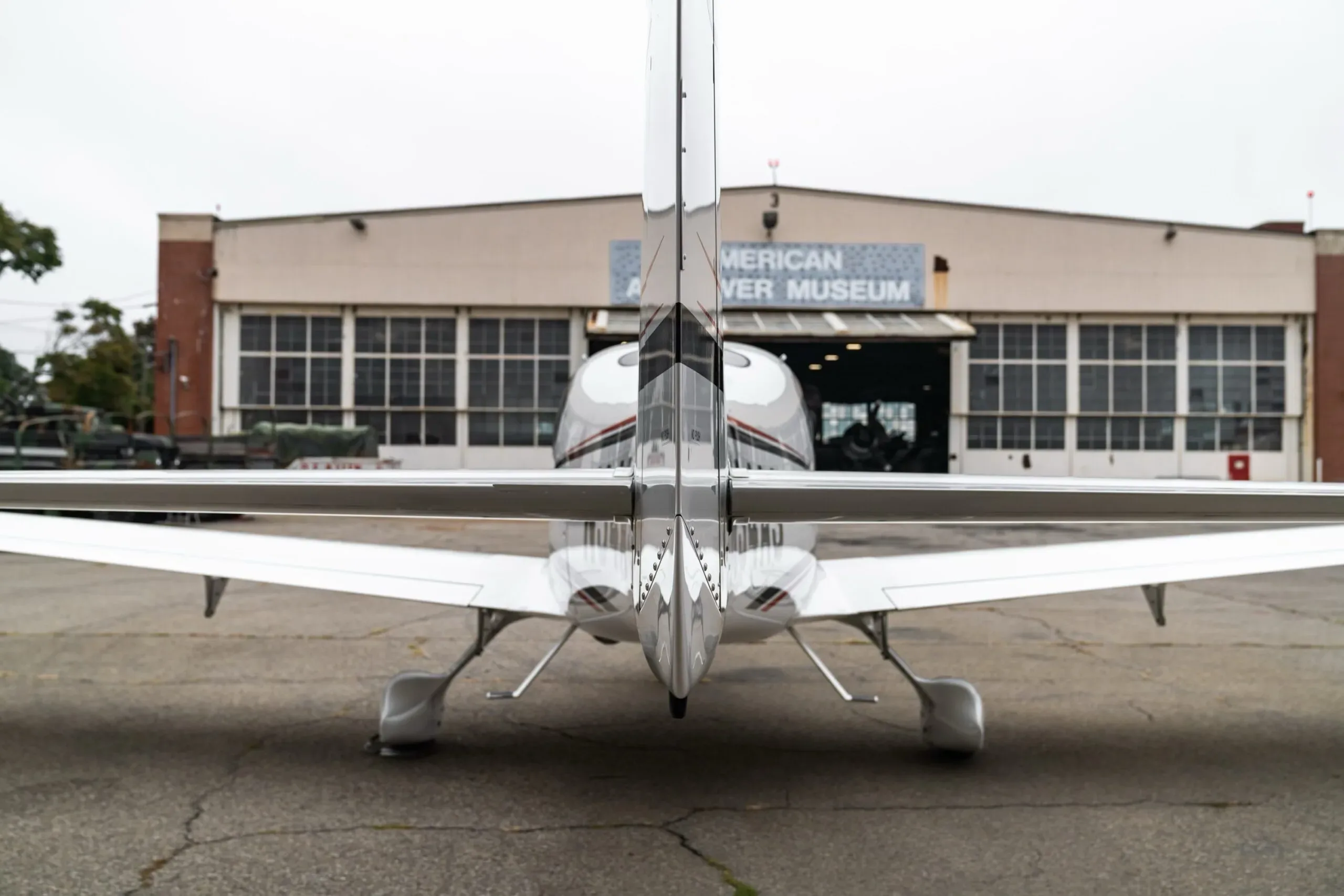 A rear view of a white Cirrus SR22 aircraft with red and black stripes, parked in front of the American Airpower Museum hangar.