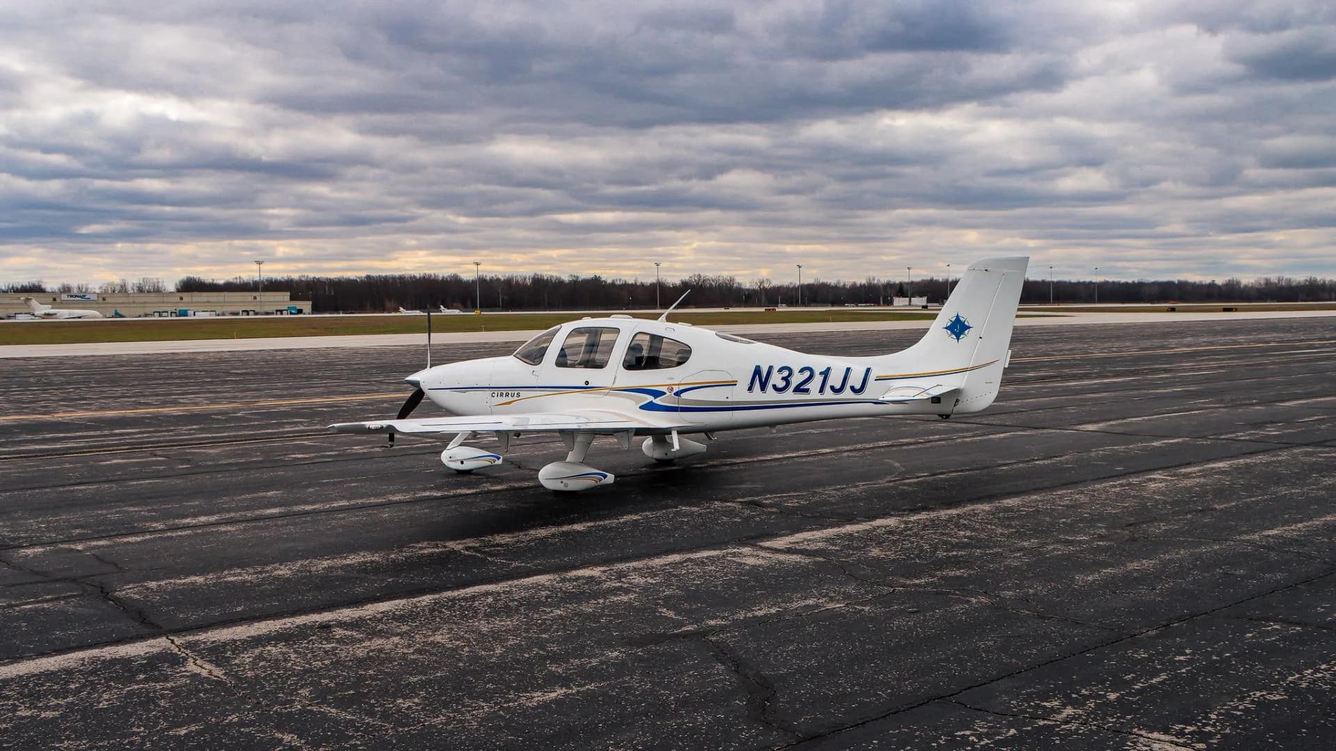 A white Cirrus aircraft, registration N321JJ, is parked on a wide airport tarmac under a cloudy sky.