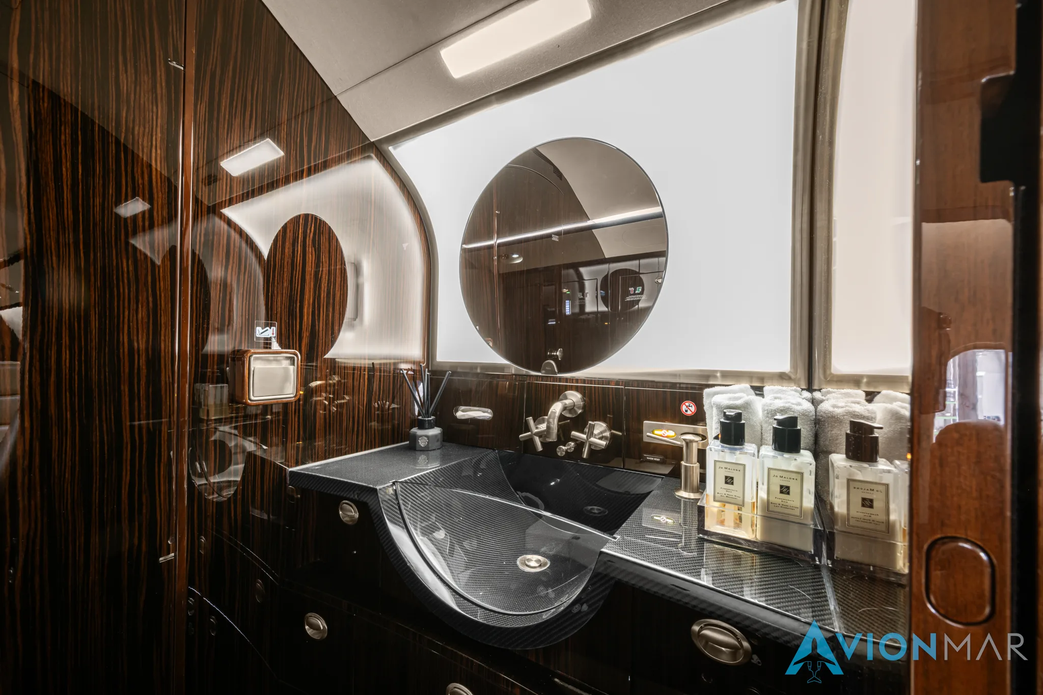 The private jet's lavatory with a carbon fiber sink, dark wood cabinetry, a round mirror, and toiletries.