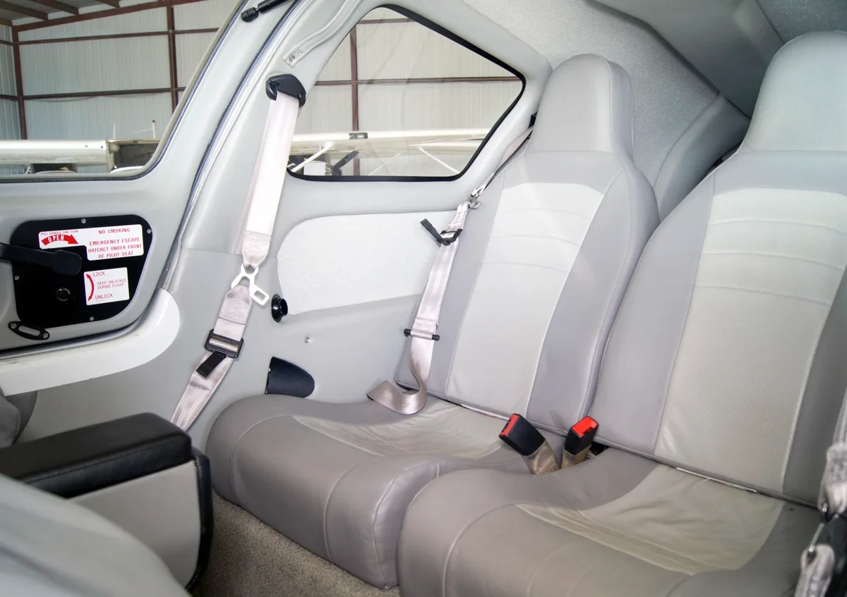 The gray leather passenger seats of the aircraft cabin are visible, with seatbelts and a window showing another aircraft outside.