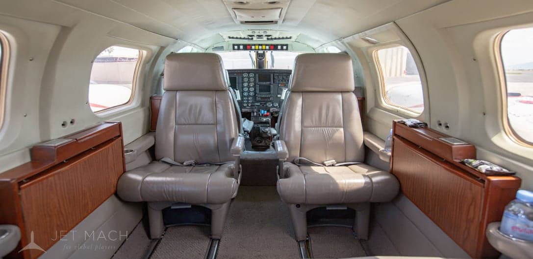 The aircraft cabin interior shows beige leather seats with seatbelts and wooden fold-out tables.