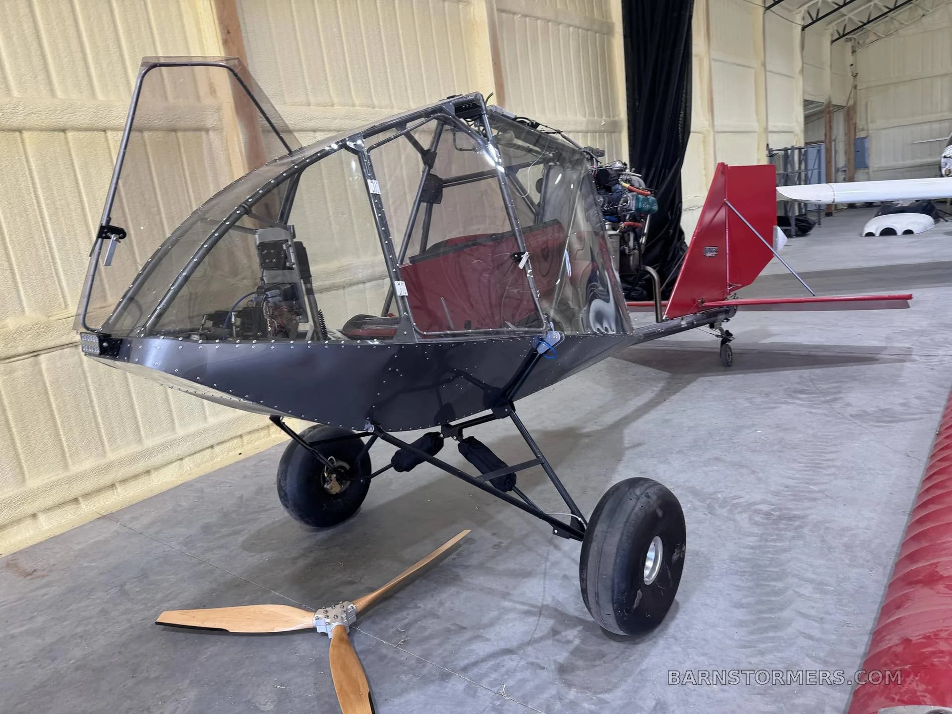 A small, open-frame ultralight aircraft with a clear bubble canopy, black fuselage, and red tail, sitting in a hangar with its propeller detached on the floor.