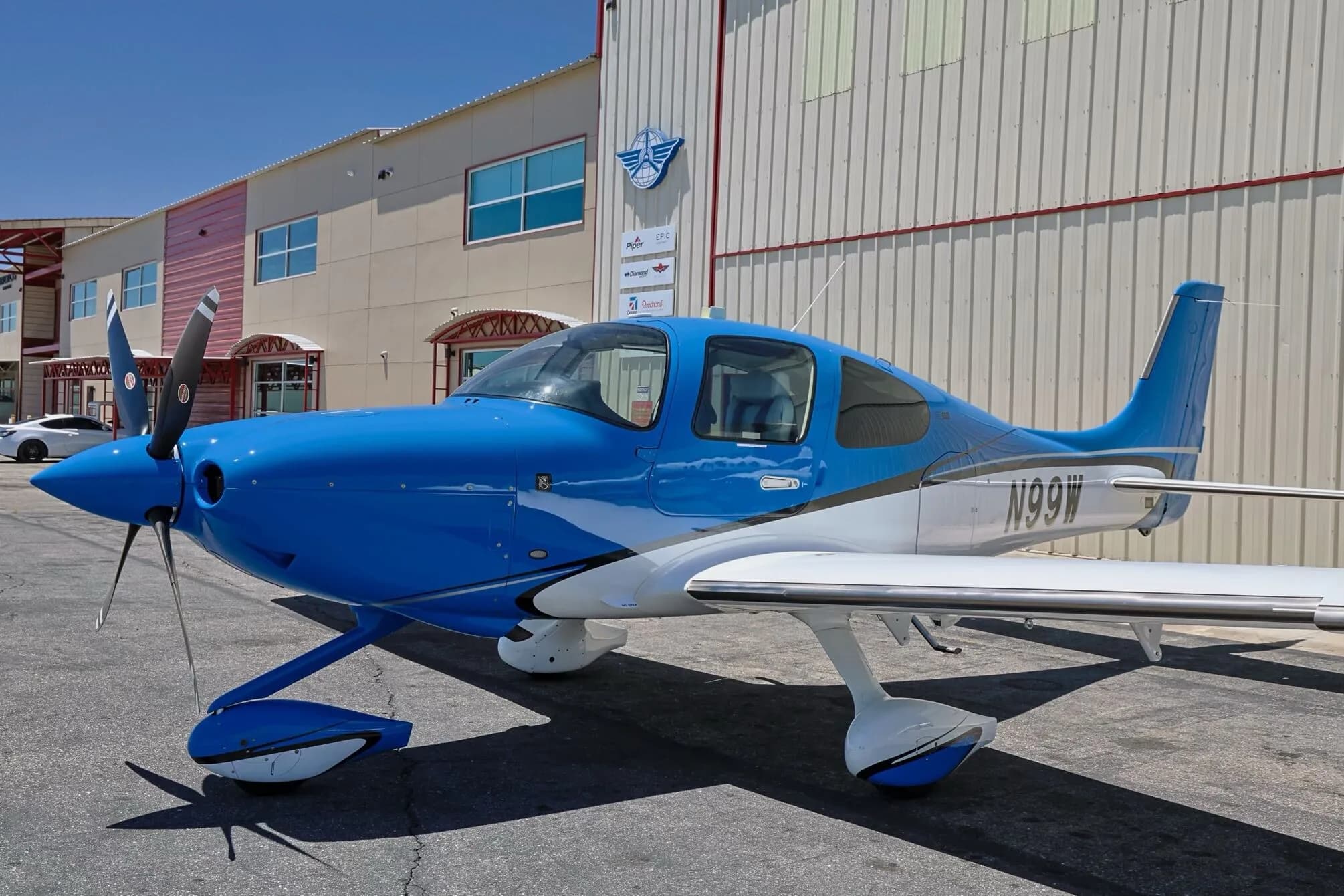 Front three-quarter view of a blue and white Cirrus SR22 aircraft (N99W) parked next to a building.