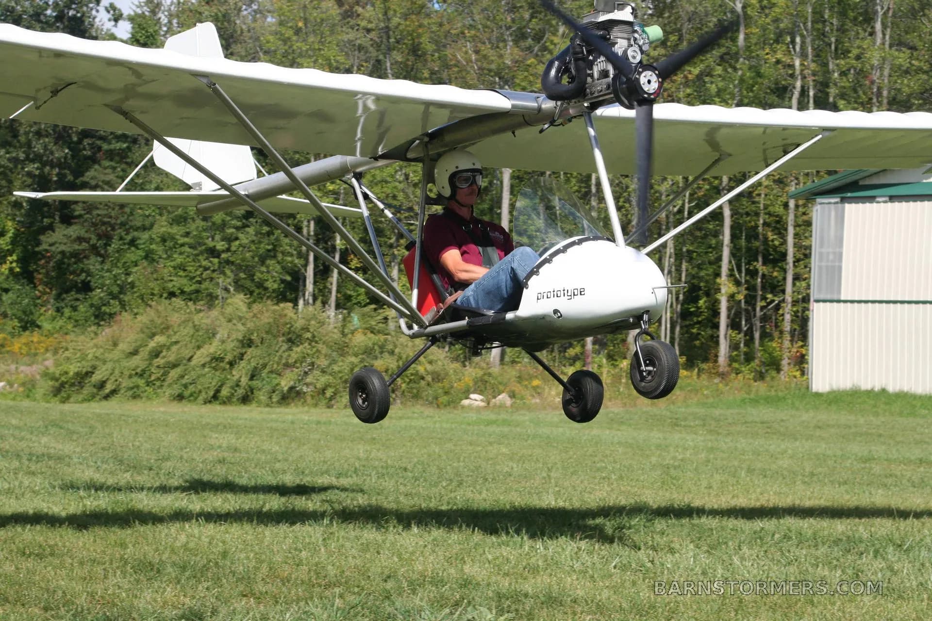 A white ultralight aircraft with 'prototype' on its nose, carrying a pilot, is shown flying low over a grassy field with trees in the background.