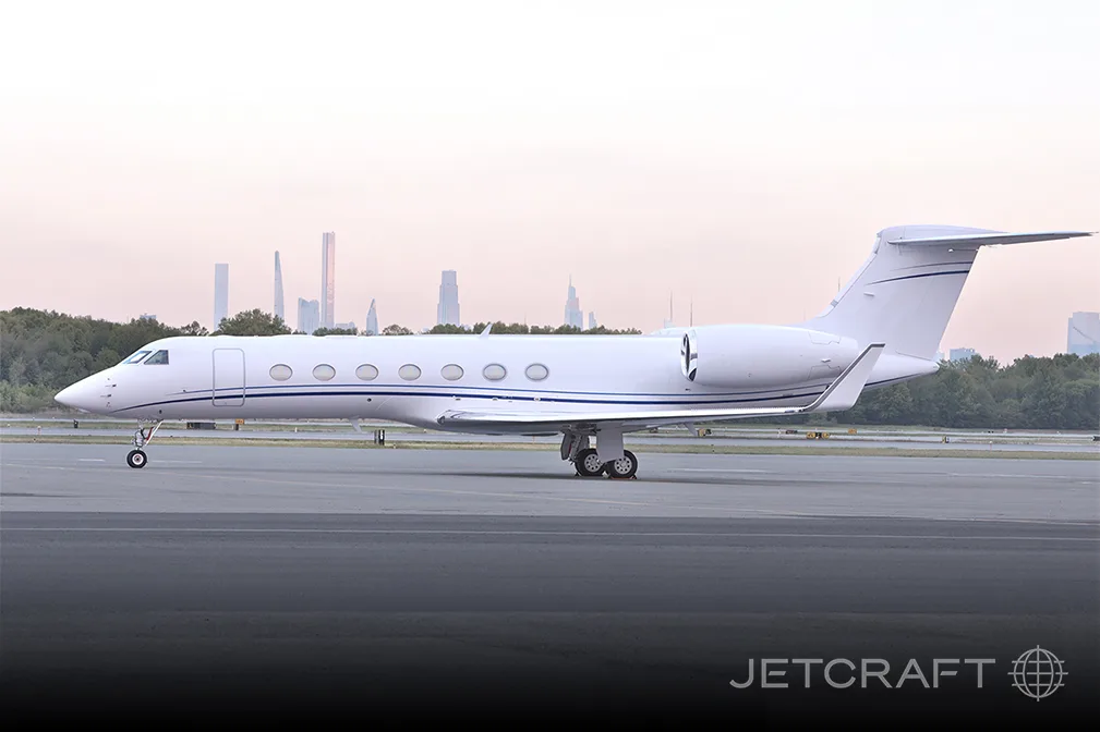 A white private jet with blue accent stripes is parked on an airport tarmac with a city skyline in the background.