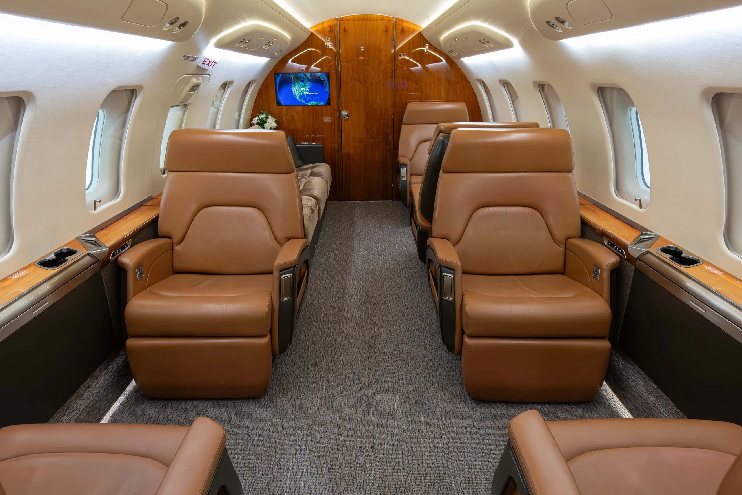 The luxurious interior of a business jet cabin features brown leather seats, wood trim, and a screen at the front.