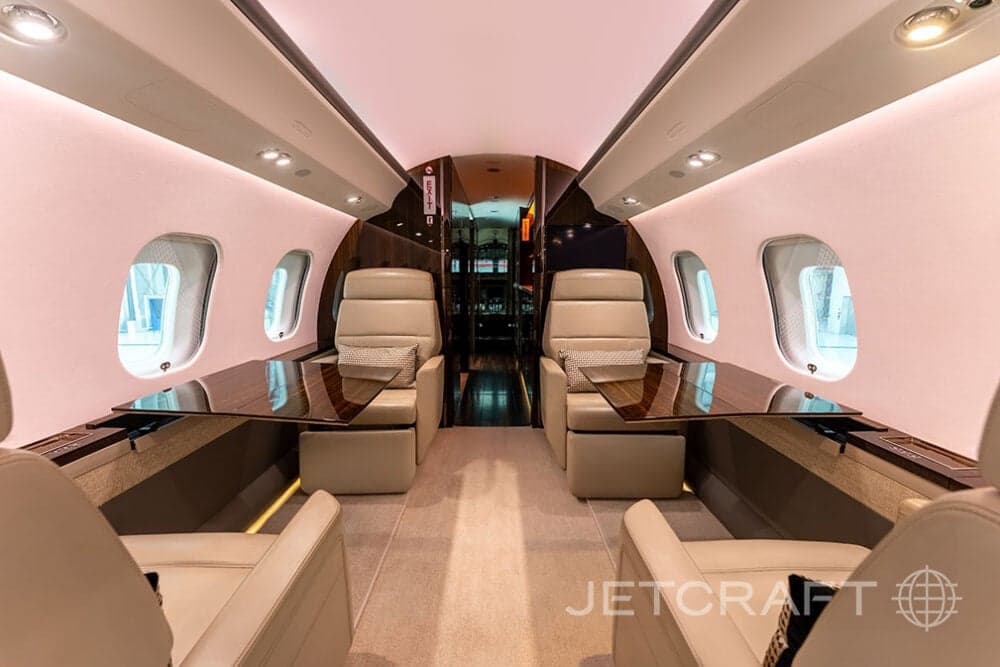 Interior view of a Bombardier Global Express business jet with beige leather seats and wooden tables.