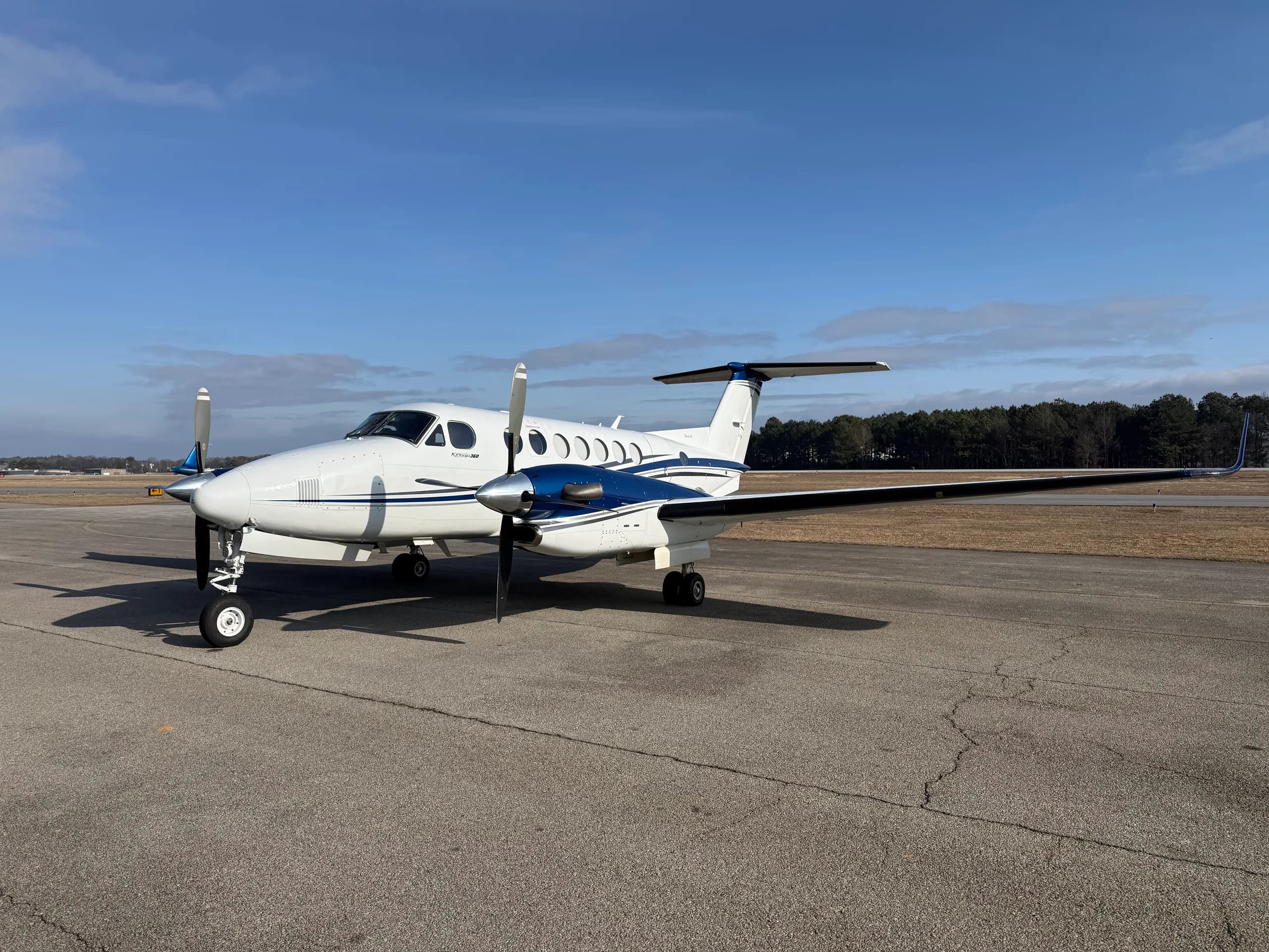 A white Beechcraft King Air 360 with blue stripes is parked on an asphalt ramp under a clear blue sky.