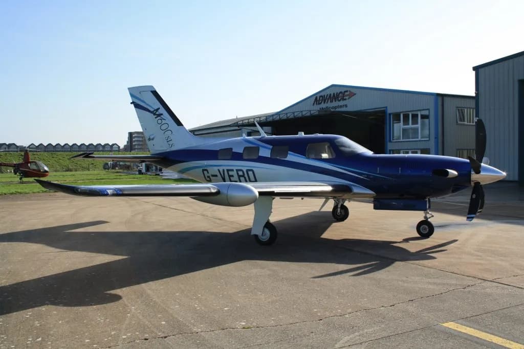 A side view of a blue and silver Piper M600SLS aircraft (G-VERO) parked on a tarmac next to an "Advance Helicopters" hangar.