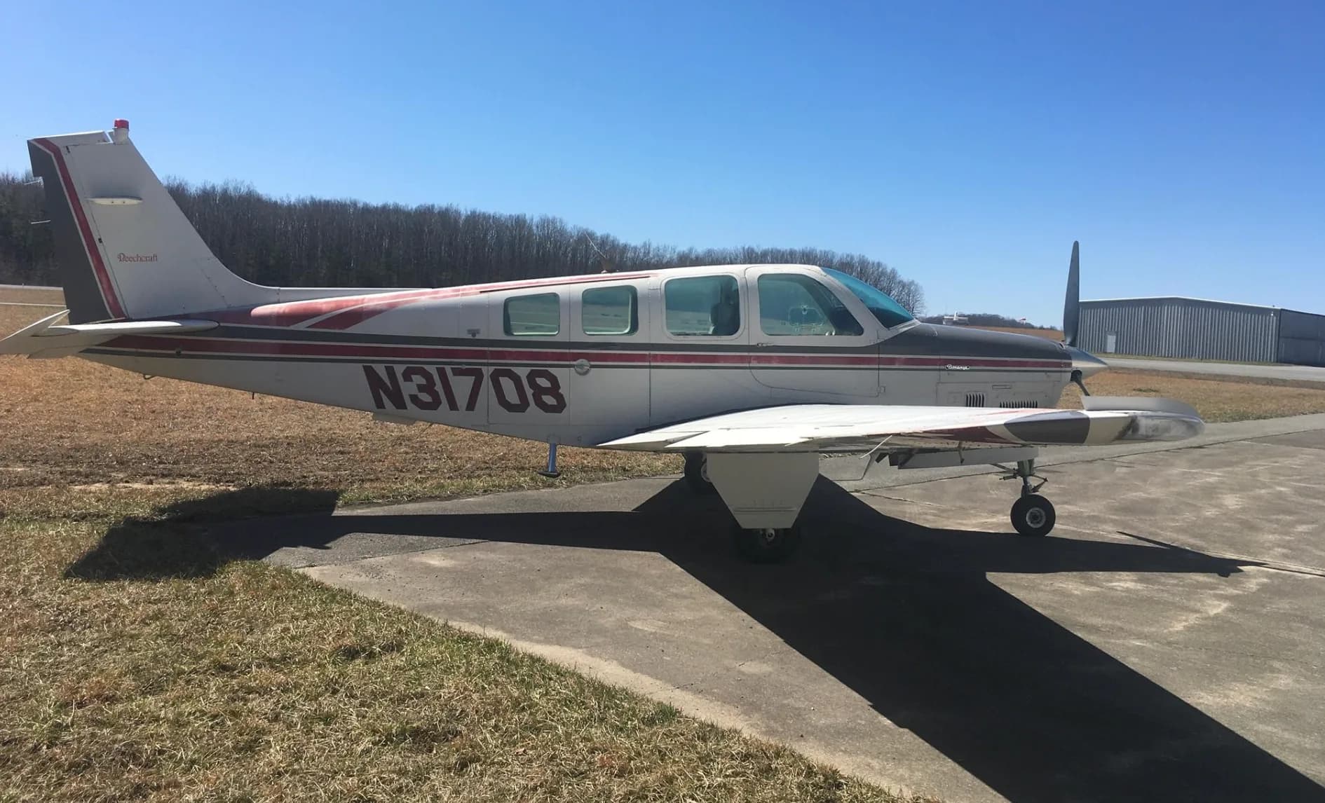 A side view of a white and red Beechcraft Bonanza N31708 parked on an asphalt ramp with a hangar and trees in the background.