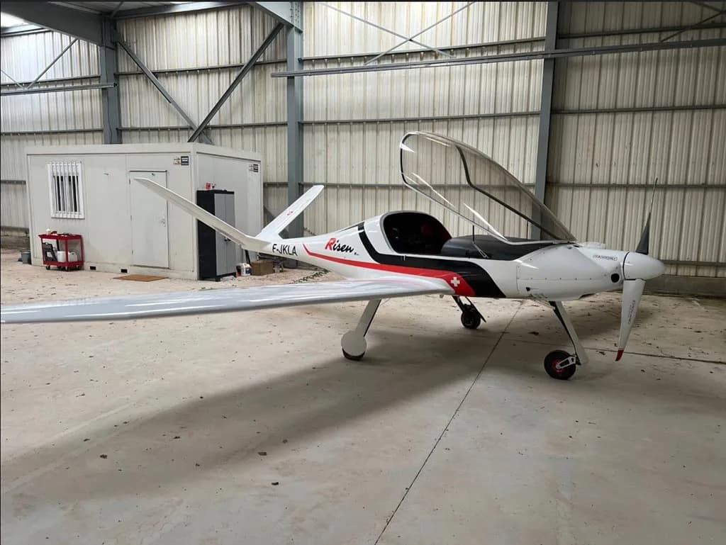 A white Risen aircraft with registration F-JKLA and an open canopy is parked inside a metal hangar.