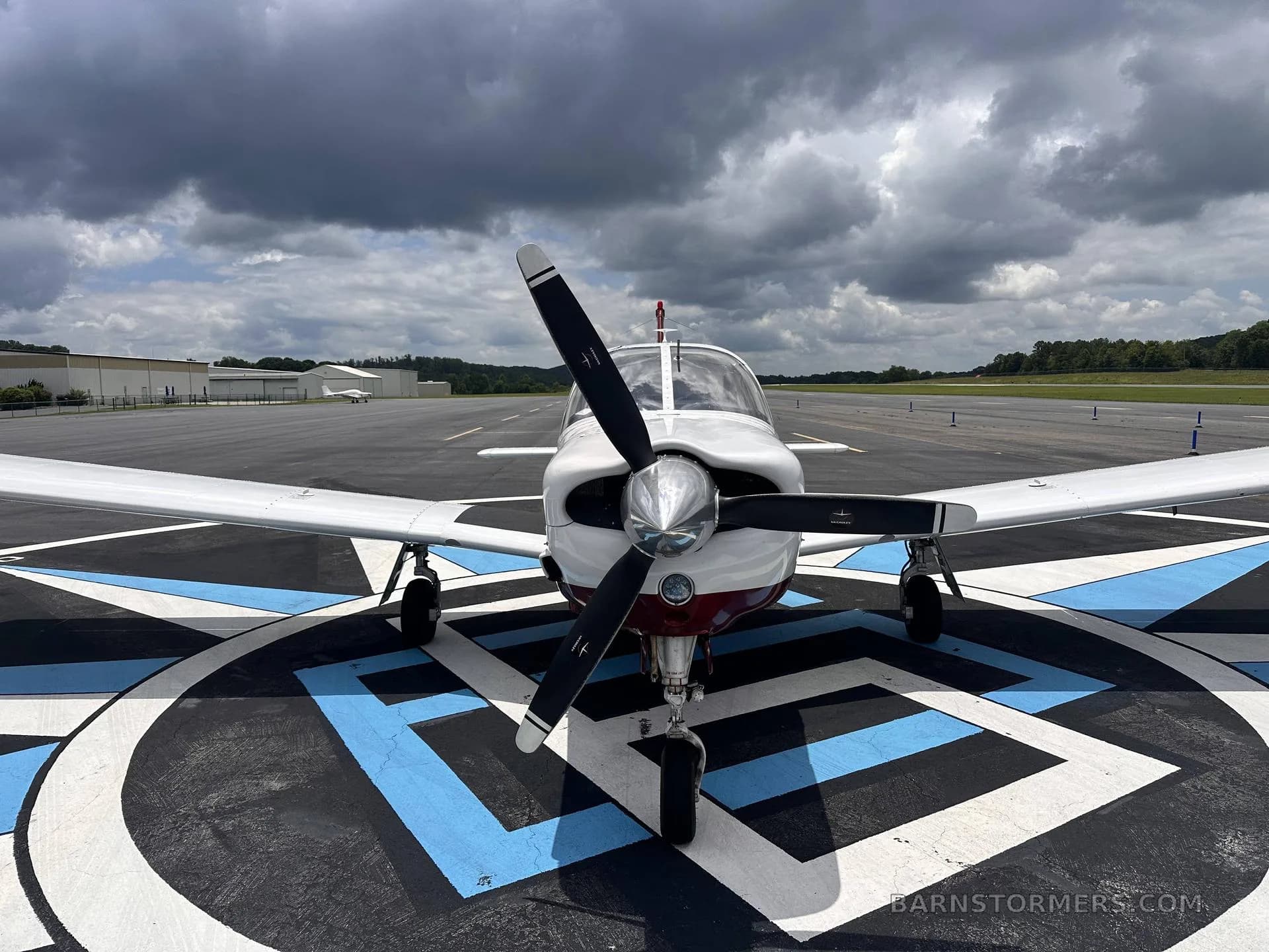 A head-on view of a white and maroon single-engine propeller aircraft (N203BW) parked on a blue and white patterned tarmac with a runway in the background.
