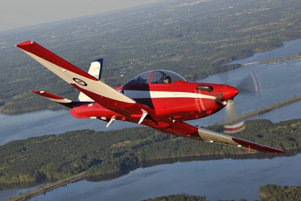 A red and white single-engine aircraft with a bubble canopy flies over a river and green landscape.