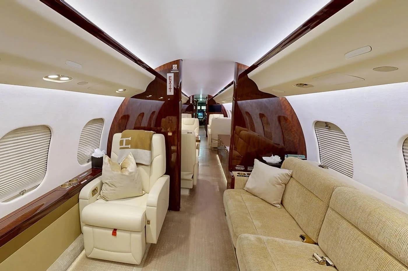 The aft section of a private jet cabin, featuring a cream-colored armchair with a blanket and a long beige sofa, all surrounded by dark wood and light walls.