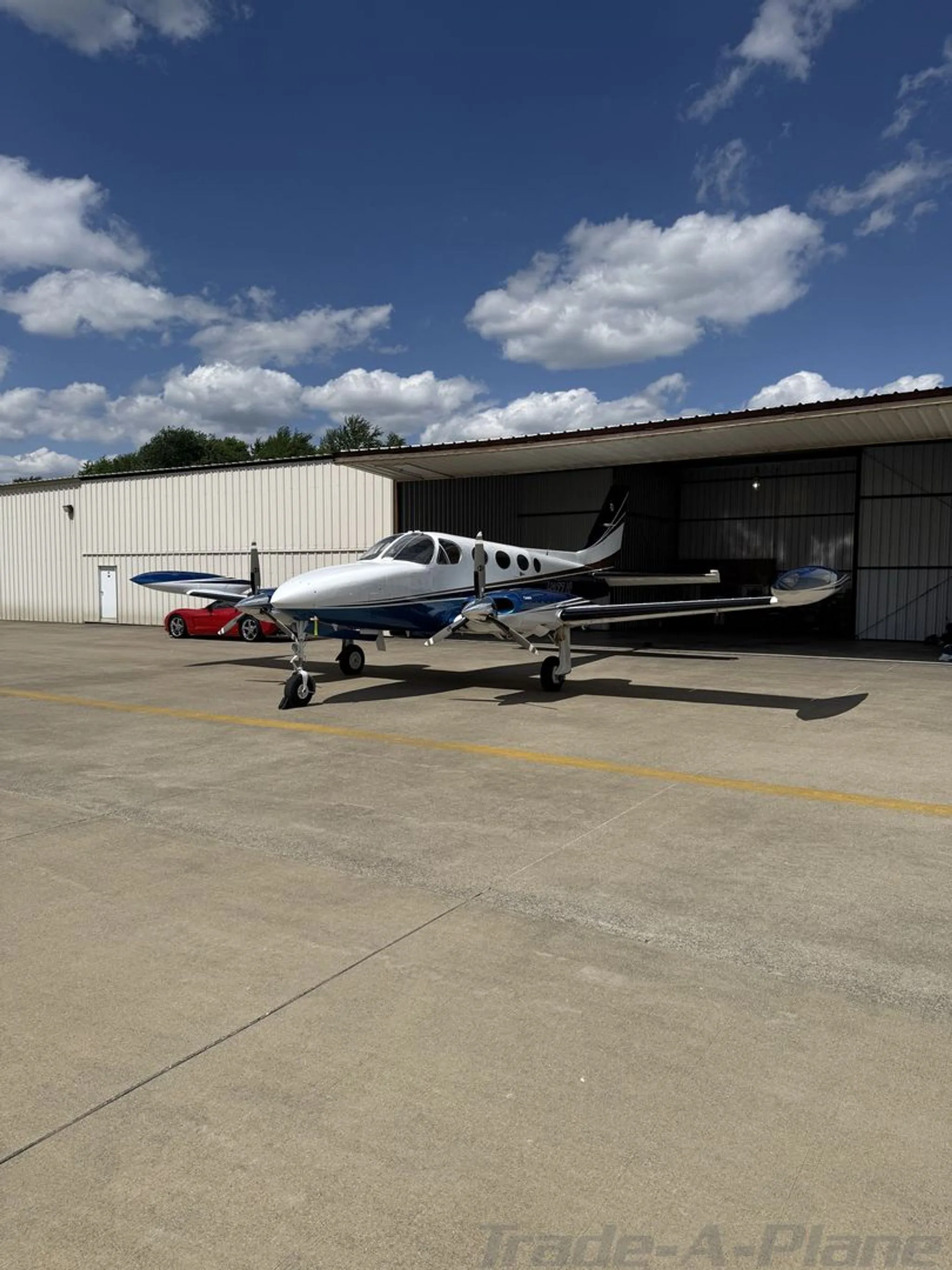 A side view of a white, blue, and gold Cessna 414 twin-engine aircraft parked on an apron next to a red car.