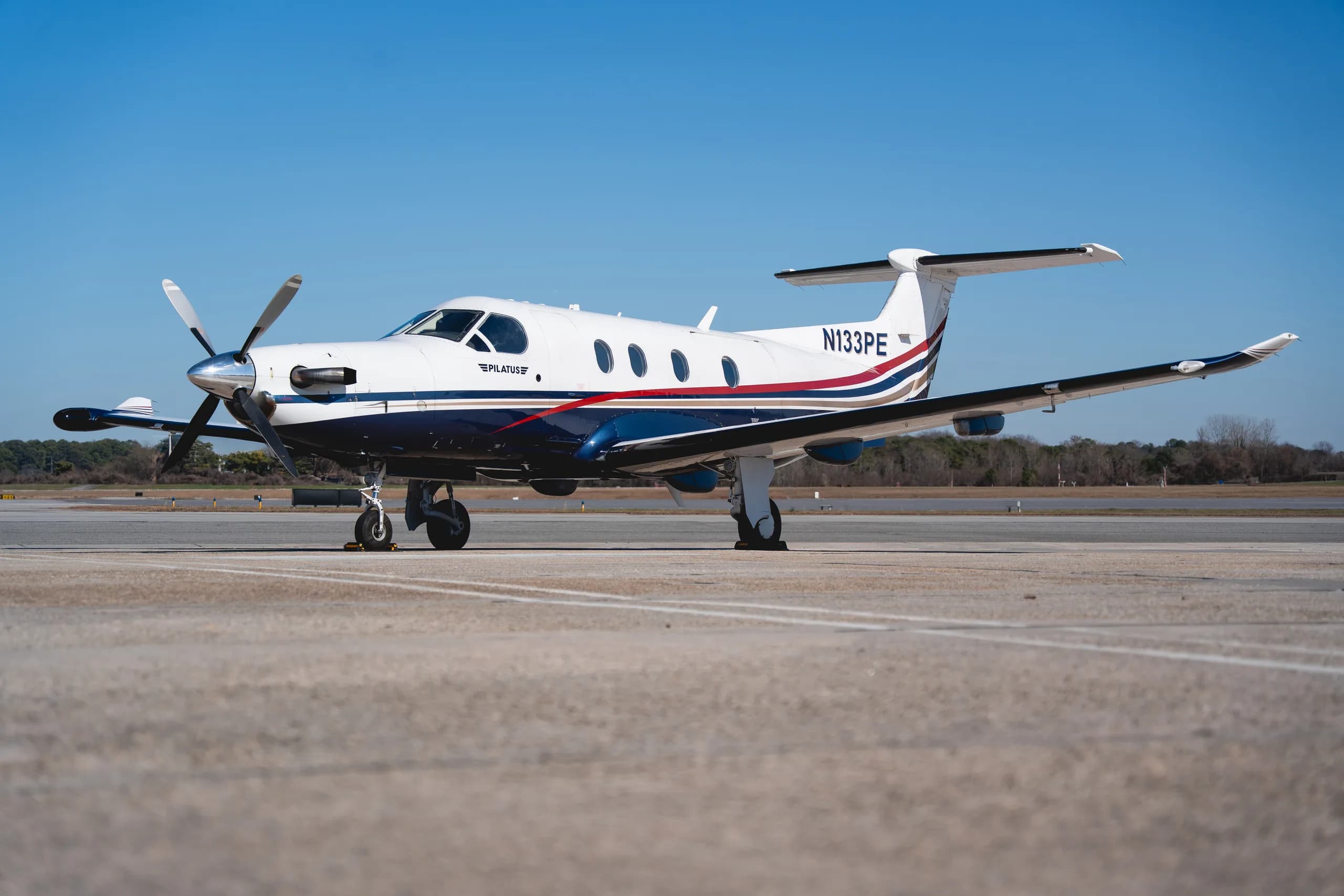 A Pilatus PC-12 turboprop aircraft, registration N133PE, with a white, blue, red, and gold stripe paint scheme, parked on an airfield under a clear blue sky.