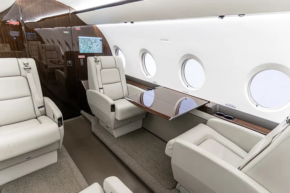 A section of a luxurious aircraft cabin showing light-colored leather seats, a fold-out table, and large windows.