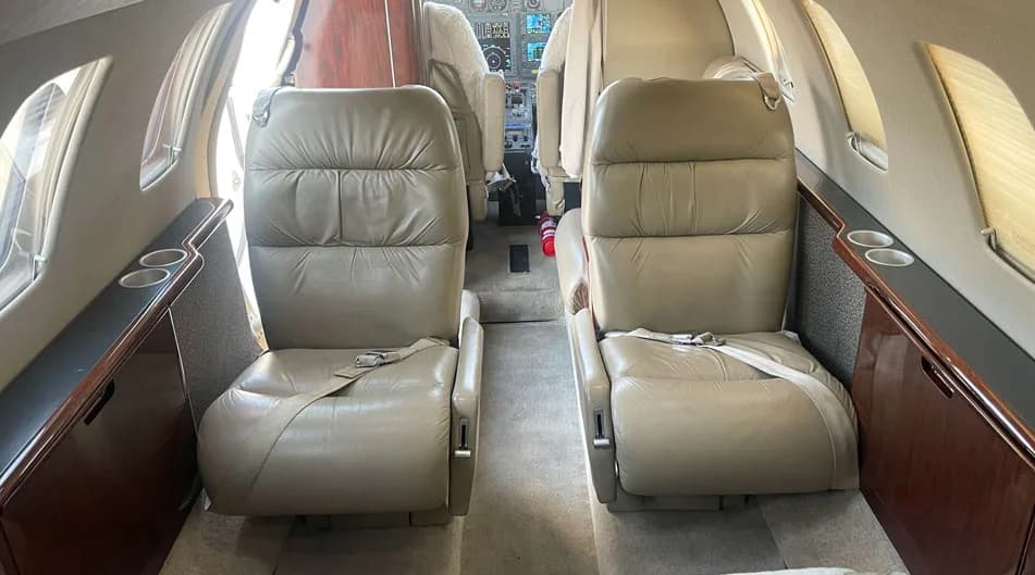 The aircraft cabin interior with two tan leather passenger seats facing forward, and a view towards the cockpit.