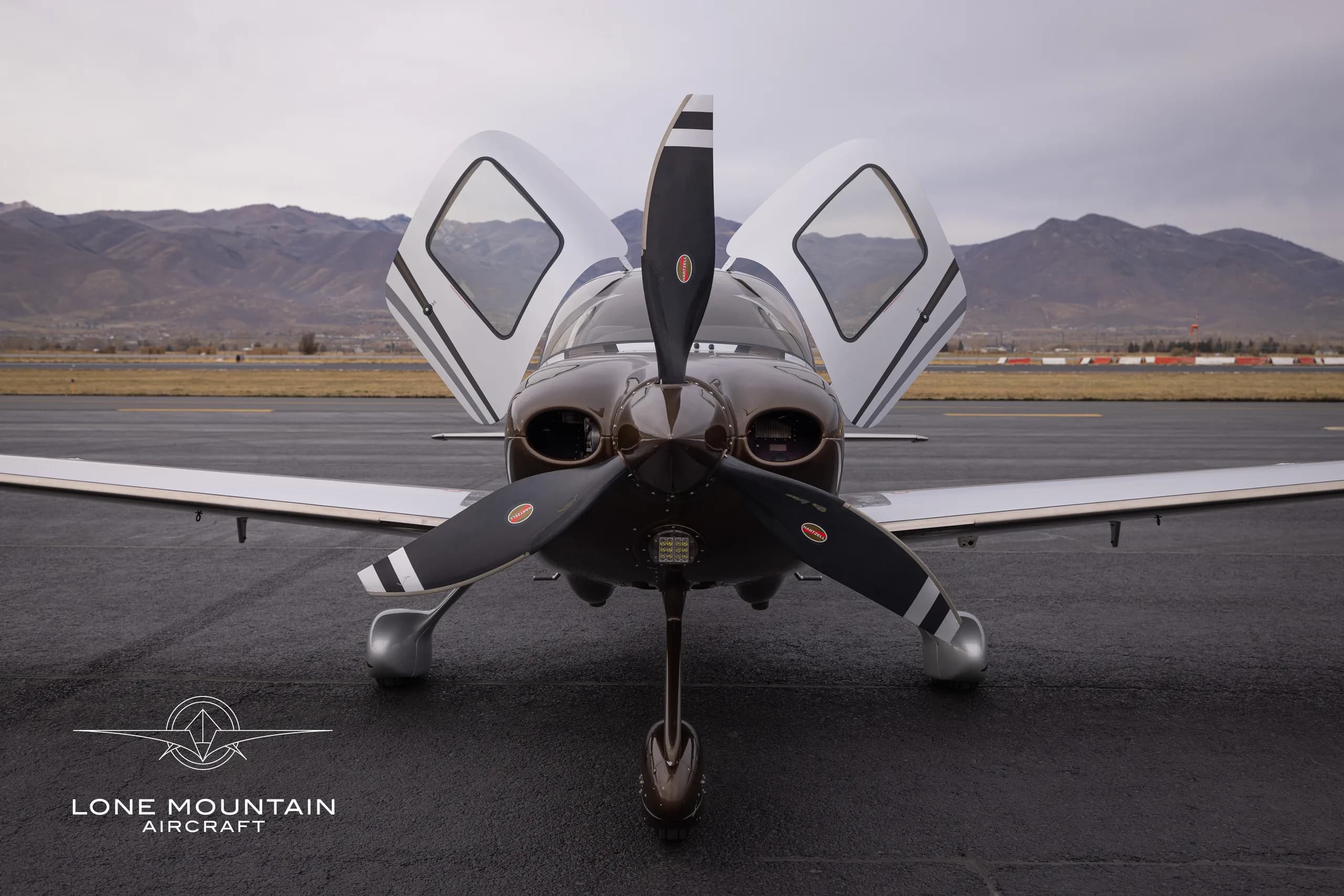 A Cirrus SR22T aircraft with a brown and white paint scheme is parked on the tarmac with the doors open, facing forward.