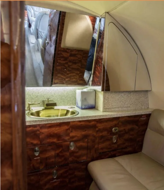 The lavatory area of a business jet, featuring a gold sink, mirror, and wood cabinetry.