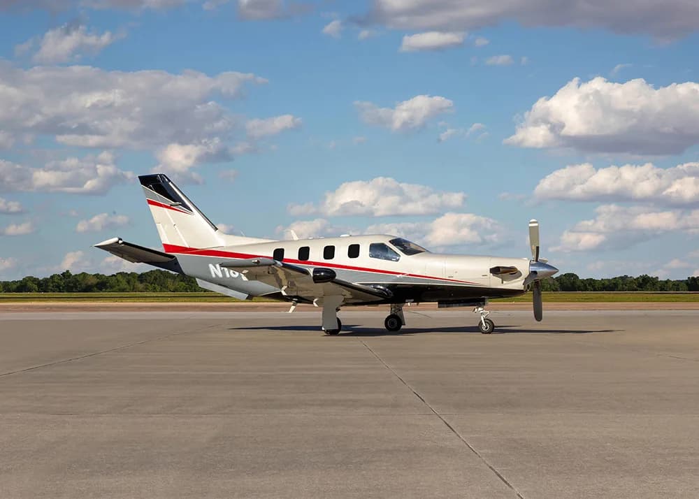 A white and grey TBM 900 series aircraft with red stripes, registration N180MF, parked on an airfield, viewed from the front-left.