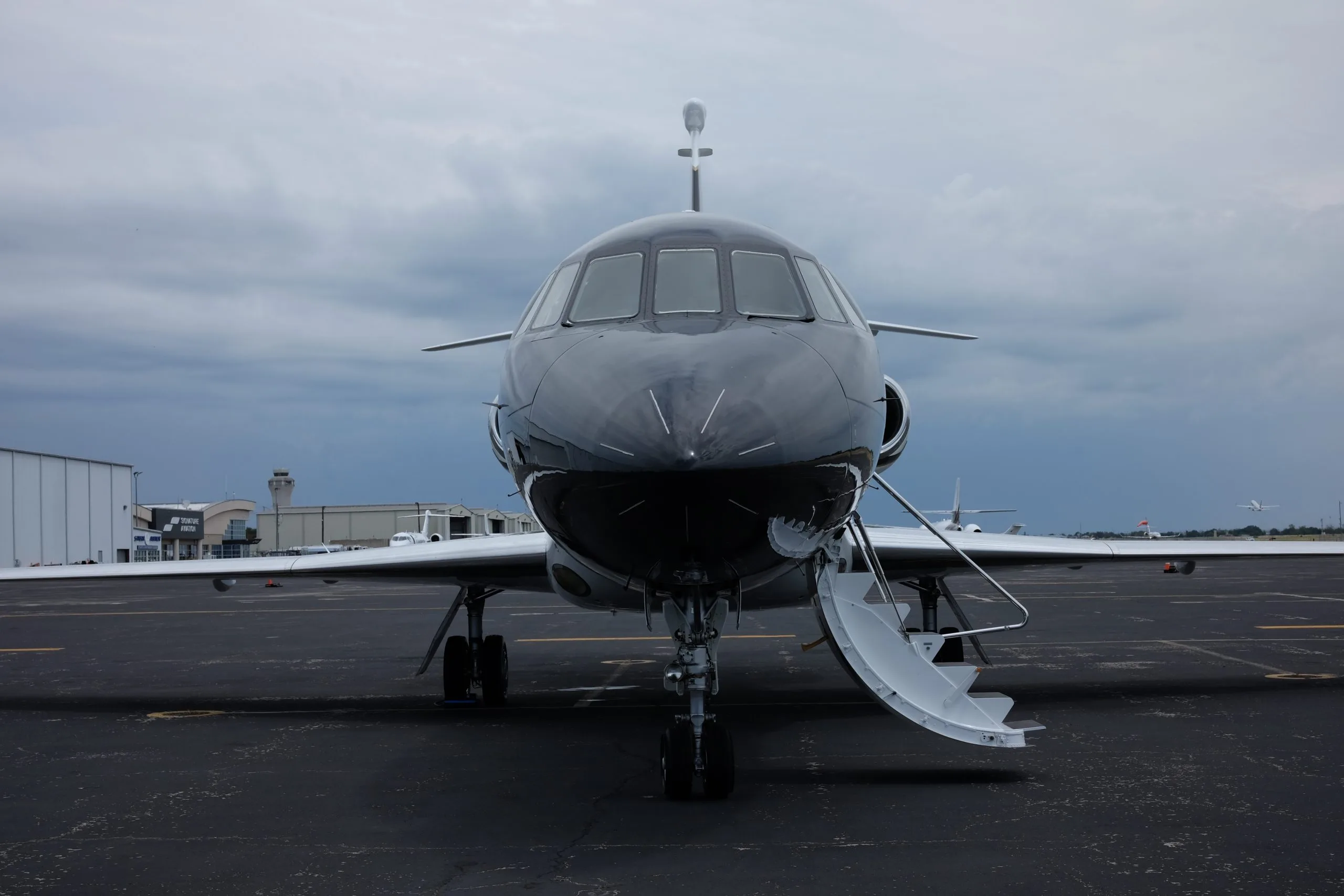 A front view of a black and white Dassault Falcon 50 business jet with its door open, parked on an airport tarmac under an overcast sky.