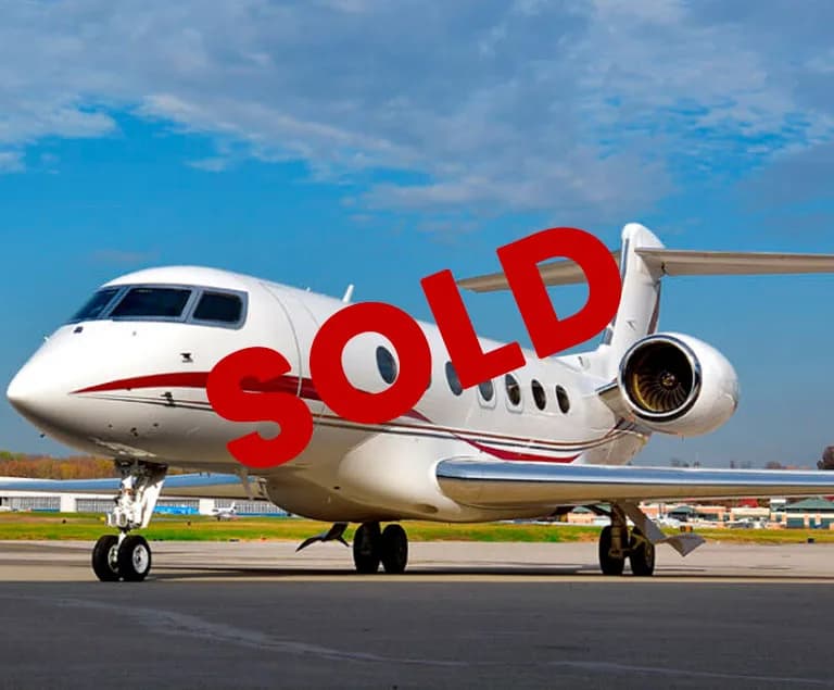 A white business jet with a red stripe on the fuselage is parked on an airfield under a blue sky.